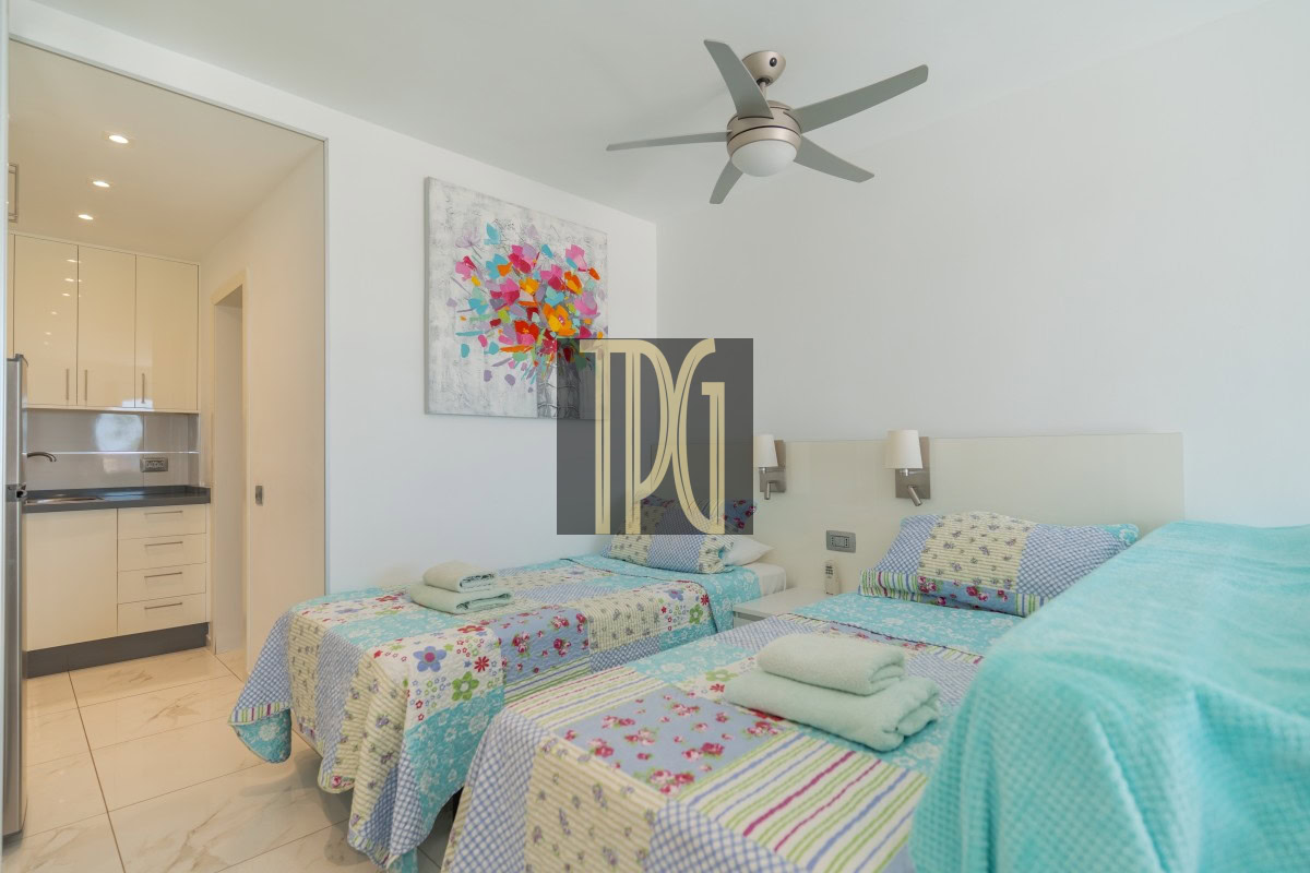 Twin beds with colorful patchwork quilts and folded towels in a bright hotel room; kitchenette visible through an open doorway and a ceiling fan overhead.