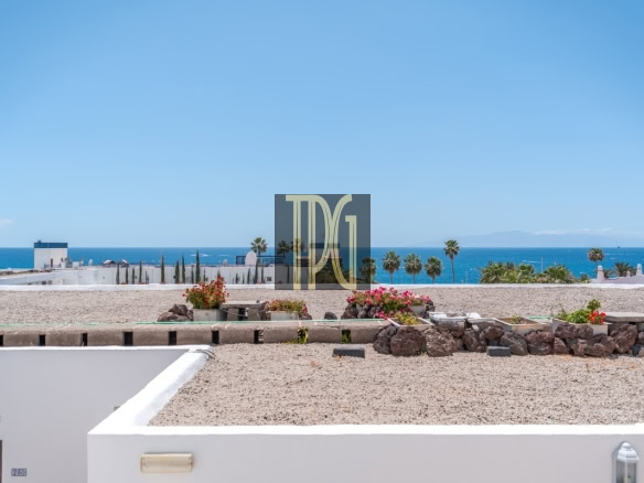 Coastal resort scene with white buildings, gravel courtyard, flowering planters, palm trees, and a blue ocean horizon.