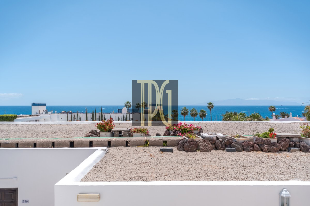 Coastal resort scene with white buildings, gravel courtyard, flowering planters, palm trees, and a blue ocean horizon.