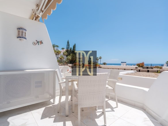 White wicker patio chairs and table on a sunlit balcony with a view of palm trees and the blue sea beyond