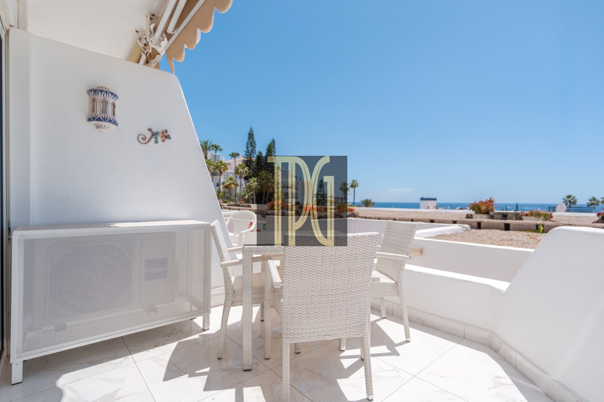 White wicker patio chairs and table on a sunlit balcony with a view of palm trees and the blue sea beyond