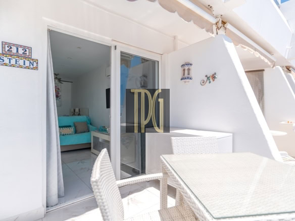 Outdoor white-washed balcony with wicker dining set and glass door opening to a turquoise-sofa living area inside (apartment interior visible).