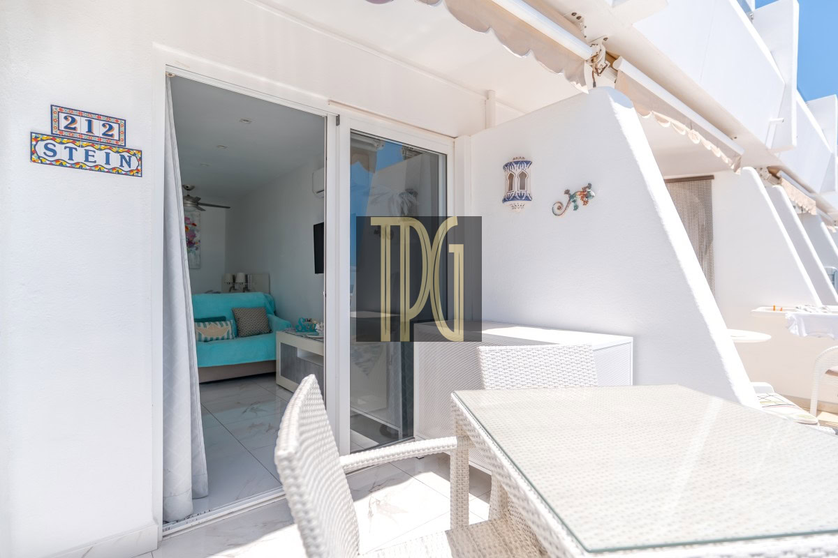 Outdoor white-washed balcony with wicker dining set and glass door opening to a turquoise-sofa living area inside (apartment interior visible).