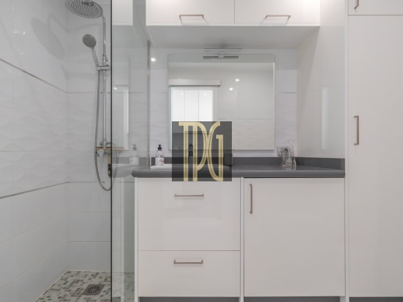 White bathroom with glass-enclosed shower on the left, gray countertop, and white cabinets beneath a mirrored vanity.