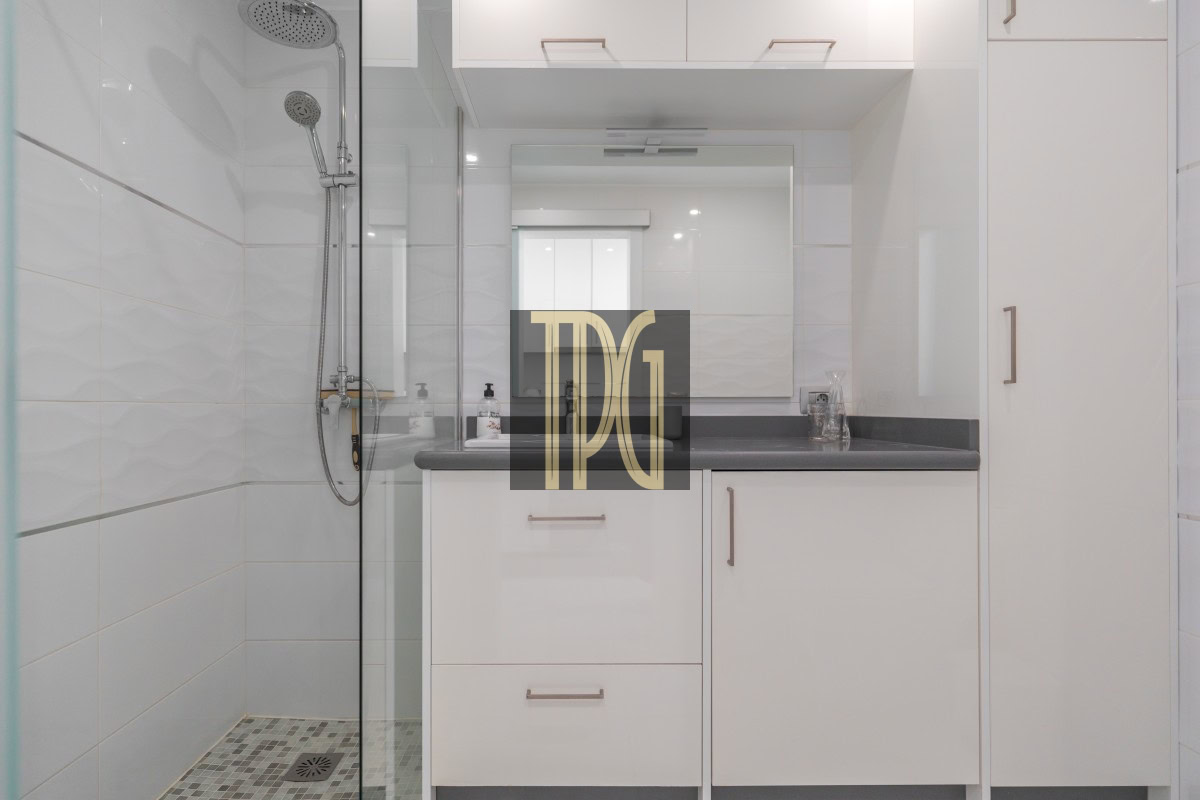 White bathroom with glass-enclosed shower on the left, gray countertop, and white cabinets beneath a mirrored vanity.