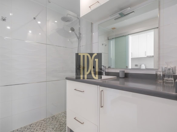 Modern bathroom with glass shower enclosure, white tiled walls, gray countertop, and white cabinetry reflected in a large mirror.