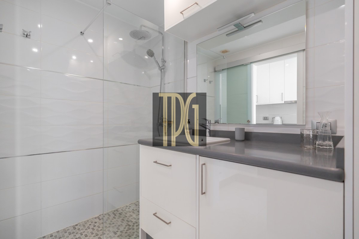 Modern bathroom with glass shower enclosure, white tiled walls, gray countertop, and white cabinetry reflected in a large mirror.