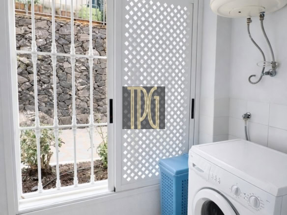 Laundry room with a front-loading washing machine, blue laundry basket, and a white lattice door beside a barred window.