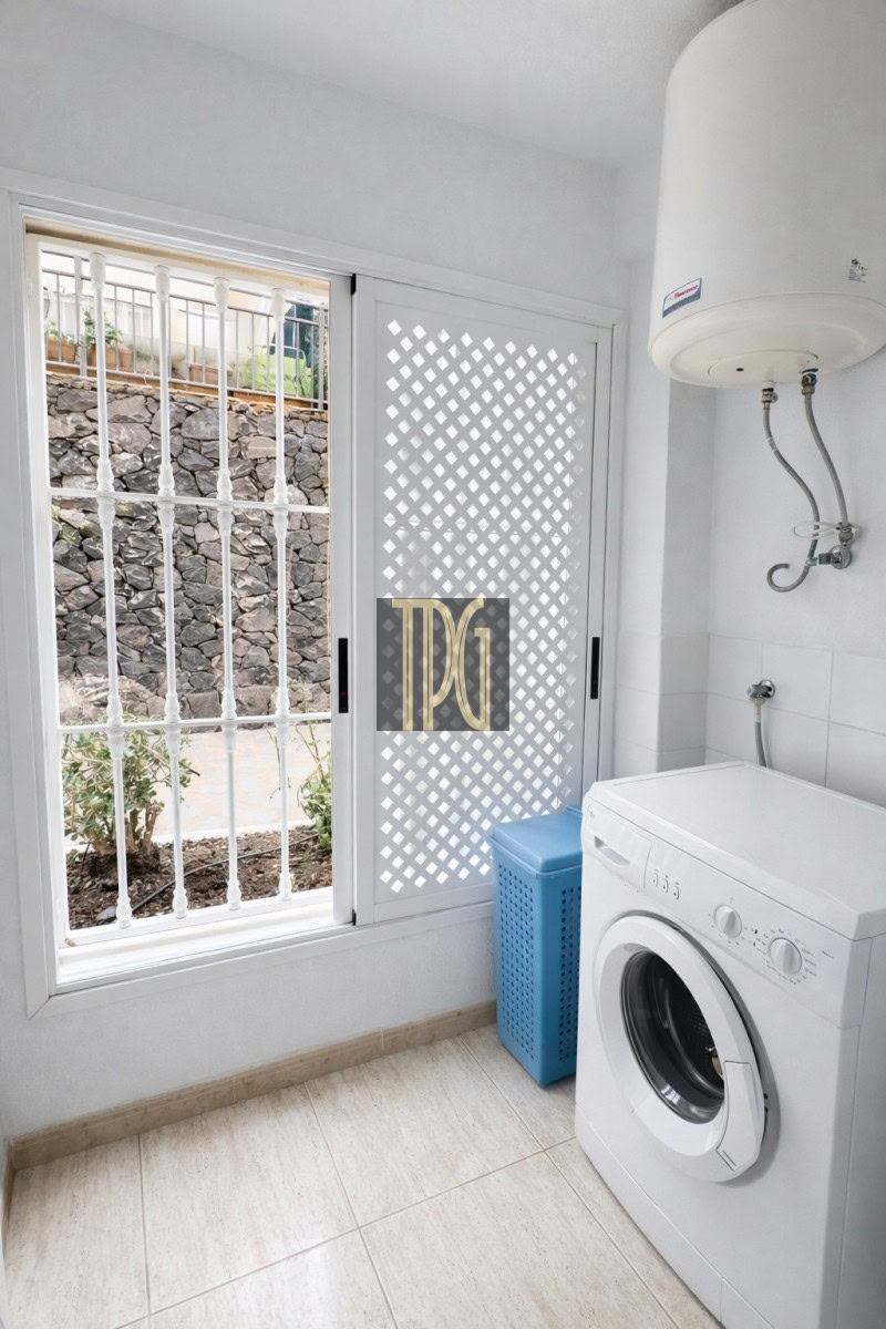 Laundry room with a front-loading washing machine, blue laundry basket, and a white lattice door beside a barred window.