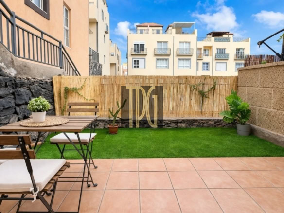 Outdoor patio with a wooden table and benches on terracotta tiles, potted plants, and a bamboo privacy fence; green lawn and beige apartment buildings in the background.