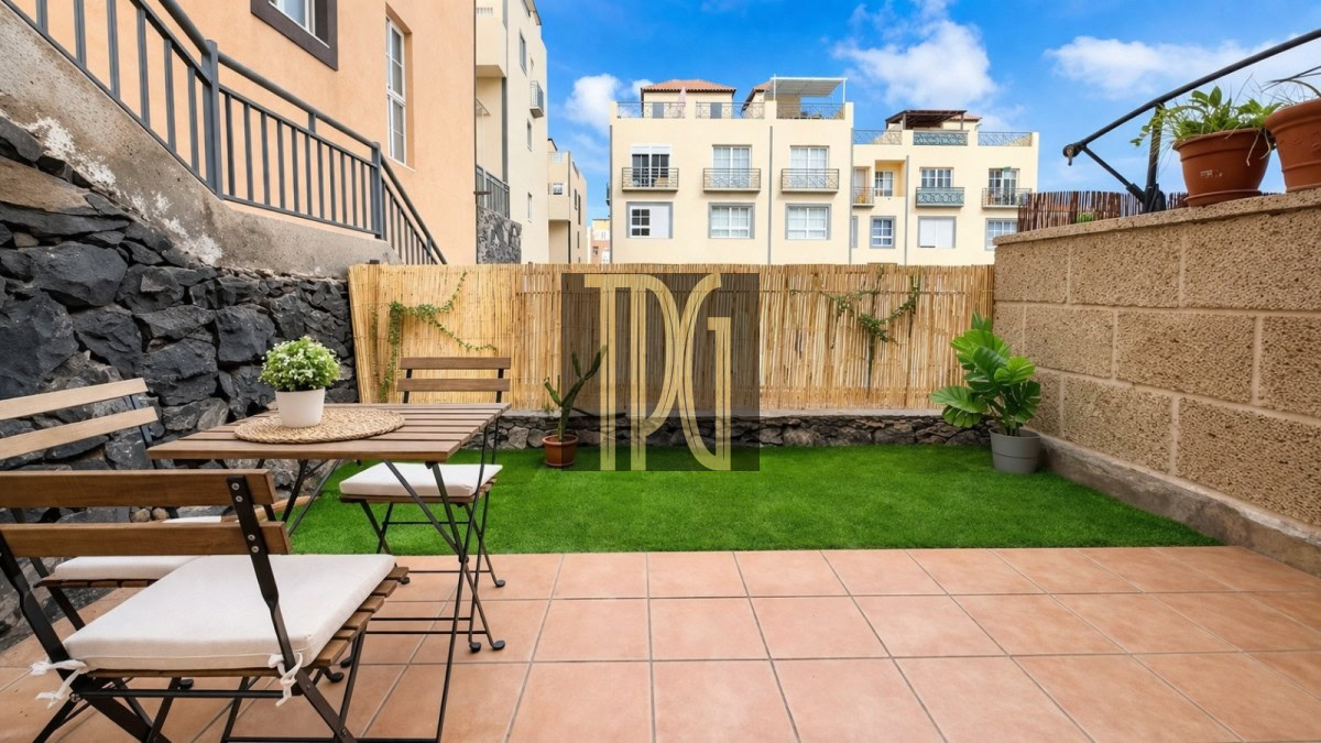 Outdoor patio with a wooden table and benches on terracotta tiles, potted plants, and a bamboo privacy fence; green lawn and beige apartment buildings in the background.