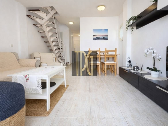 Bright living room with beige sofa, white coffee table, and a wooden dining table in the back; spiral stairs along the left wall.