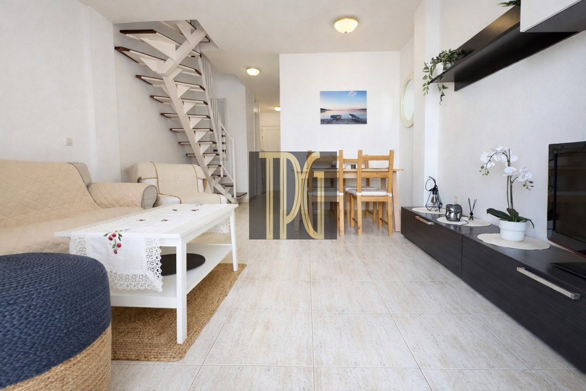 Bright living room with beige sofa, white coffee table, and a wooden dining table in the back; spiral stairs along the left wall.