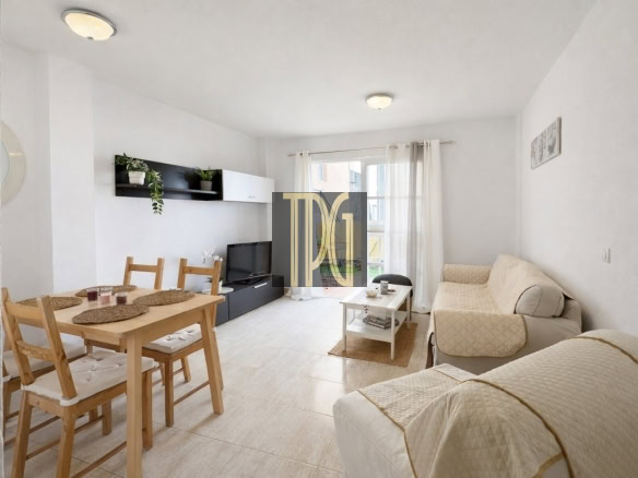 Bright living room with beige sofa and armchair, white curtains, a wooden dining table for four, a low black TV stand, and wall shelf with plants under natural light from a window.