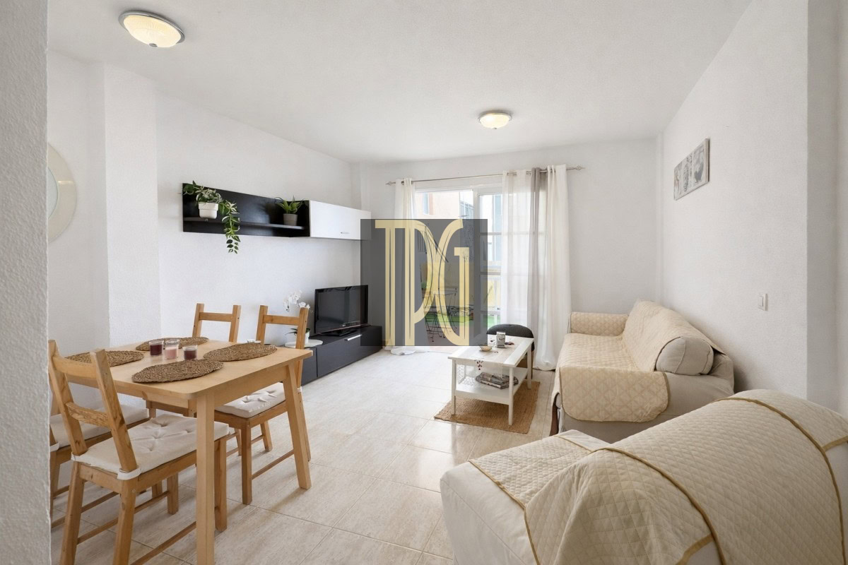 Bright living room with beige sofa and armchair, white curtains, a wooden dining table for four, a low black TV stand, and wall shelf with plants under natural light from a window.