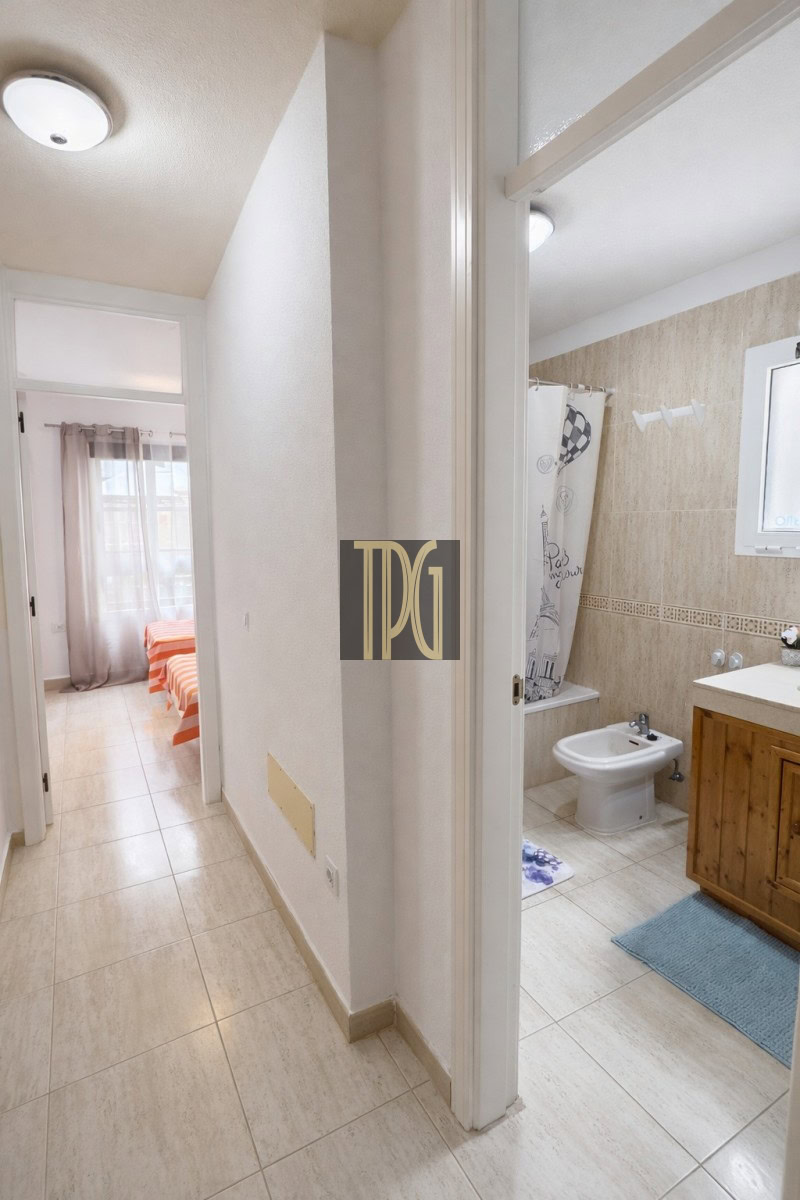 Narrow hallway with beige tiled floor; open bathroom on the right with a sink and bidet, and a bedroom with an orange-striped bed at the far end.
