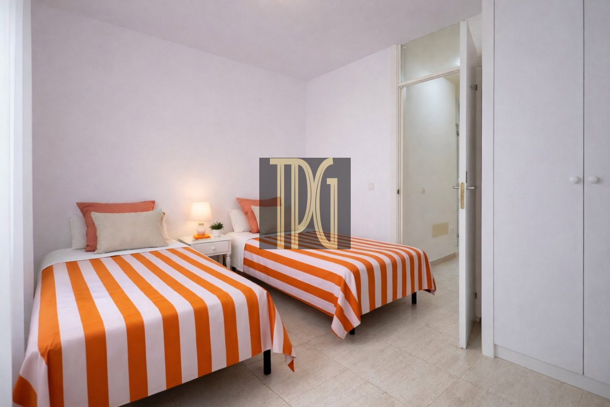 Twin bedroom with two single beds, orange-and-white striped duvets, white pillows, a small bedside table with a lamp and plant, and a closet nearby.