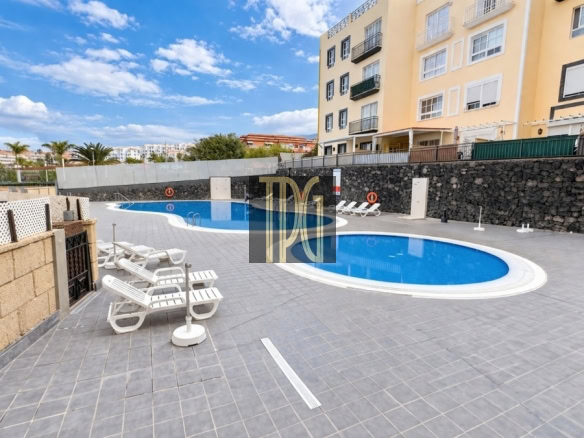 Outdoor communal swimming pool area with a curved blue pool, gray tiled deck, and white lounge chairs under a bright, sunny sky; beige apartment buildings overlook the pool.