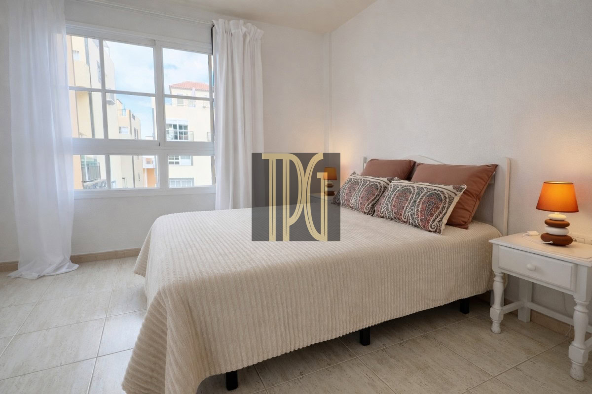 Bright bedroom with a white bedspread, decorative pillows, a white nightstand, and an orange-lamped lamp near the bed.