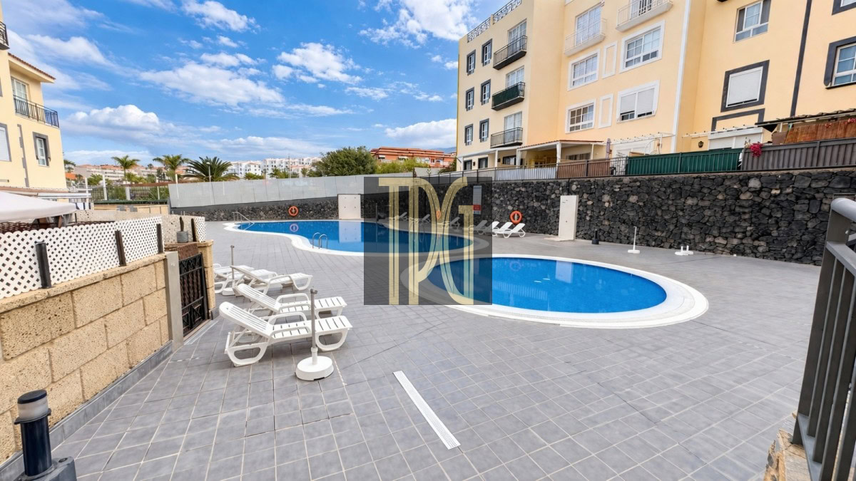Outdoor communal swimming pool area with a curved blue pool, gray tiled deck, and white lounge chairs under a bright, sunny sky; beige apartment buildings overlook the pool.