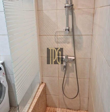 Shower area with a glass panel, handheld shower on a vertical rail, tiled walls and floor, and a drain in the center.