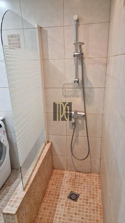 Shower area with a glass panel, handheld shower on a vertical rail, tiled walls and floor, and a drain in the center.