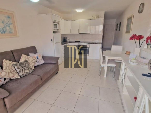 Open-plan living area with a gray sofa, patterned cushions, white tile floor, and a white kitchen in the background.