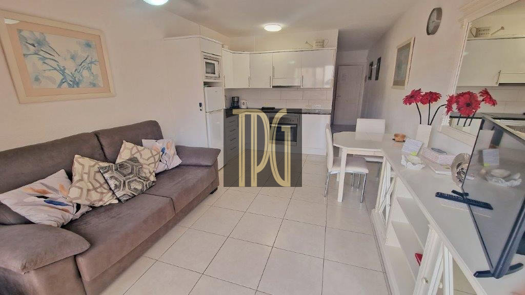Open-plan living area with a gray sofa, patterned cushions, white tile floor, and a white kitchen in the background.