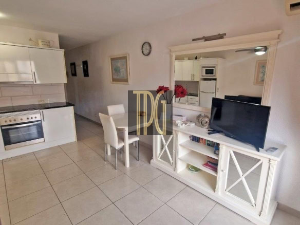 Open-plan living area with a white kitchen, dining table, and a TV on a white console under a large mirror.