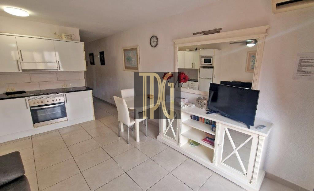 Open-plan living area with a white kitchen, dining table, and a TV on a white console under a large mirror.