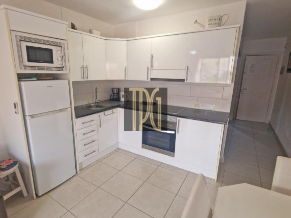 White L-shaped kitchen with fridge, microwave, sink, and built-in oven; glossy cabinets and tiled floor nearby.