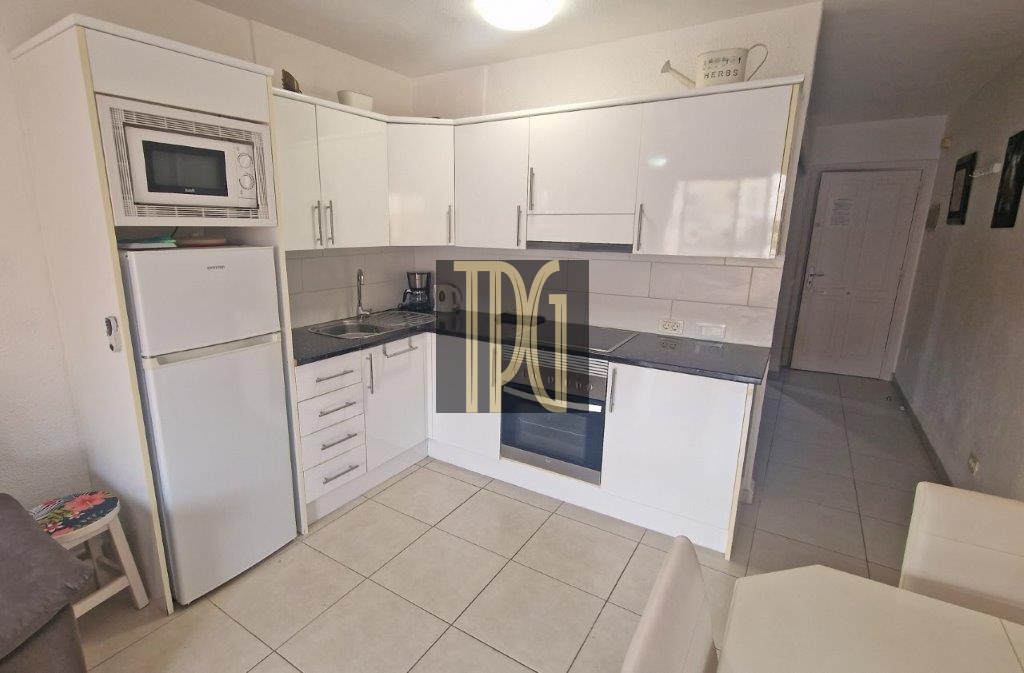 White L-shaped kitchen with fridge, microwave, sink, and built-in oven; glossy cabinets and tiled floor nearby.