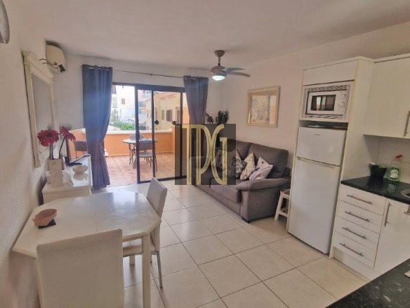 Open-plan living space with a white dining table, white chairs, a gray sofa, and a compact kitchen; blue-curtained balcony doors ahead.