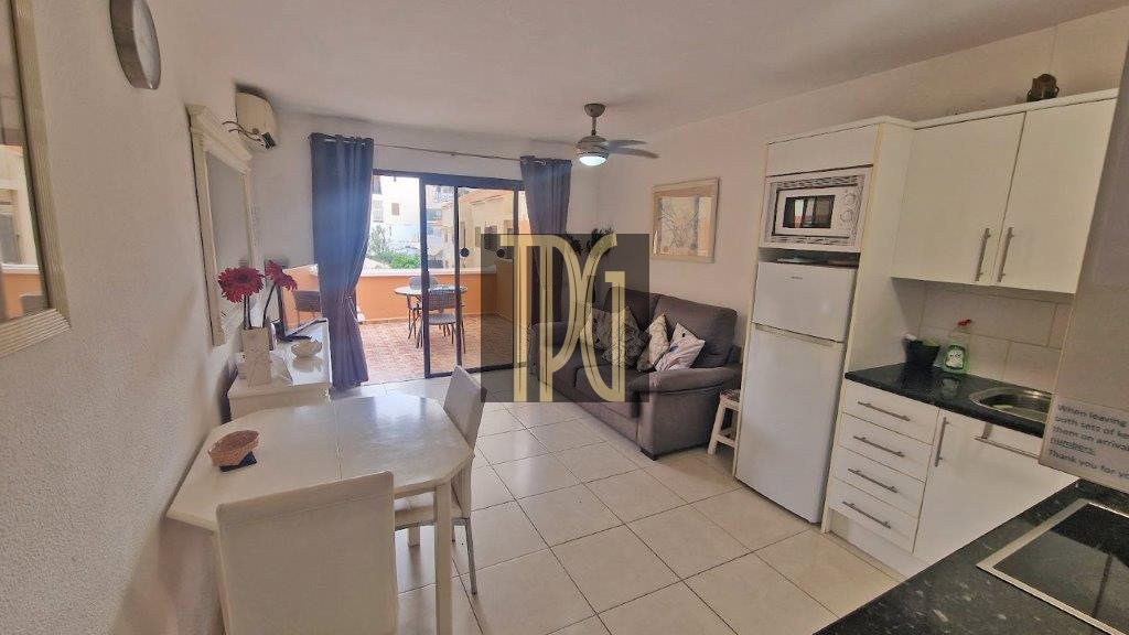 Open-plan living space with a white dining table, white chairs, a gray sofa, and a compact kitchen; blue-curtained balcony doors ahead.