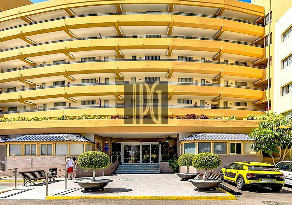 Entrance to a large yellow apartment building with curved balconies and glass doors, cars parked in front.