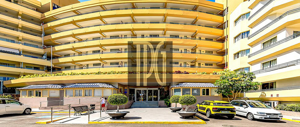 Entrance to a large yellow apartment building with curved balconies and glass doors, cars parked in front.