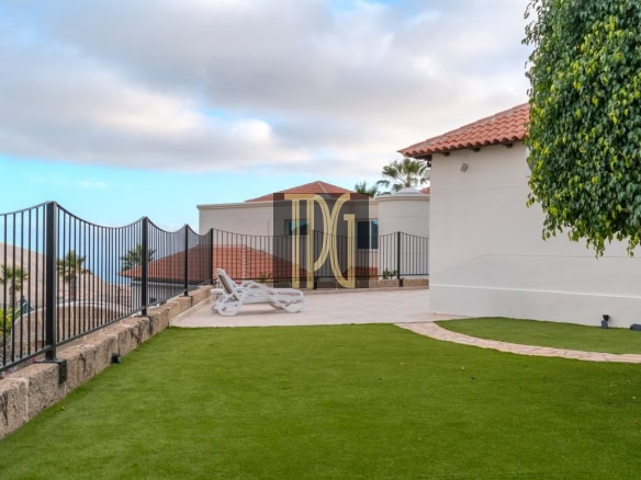 Patio with two white lounge chairs, curved stone path, green lawn, and a white building with a red-tiled roof by the sea.