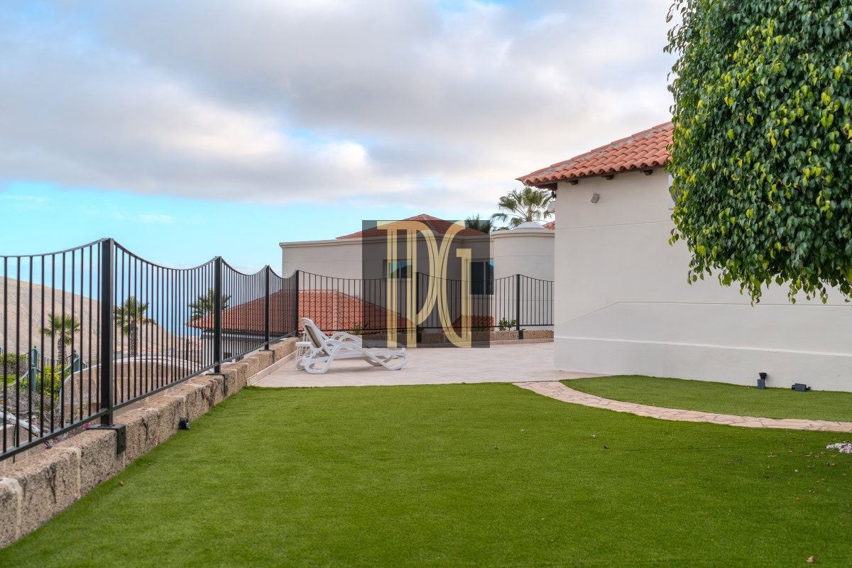 Patio with two white lounge chairs, curved stone path, green lawn, and a white building with a red-tiled roof by the sea.