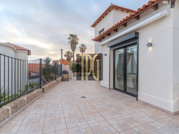 Terrace of a modern villa with sliding glass doors, tiled patio, and a black iron fence; palm trees and planters along the edge in the distance.