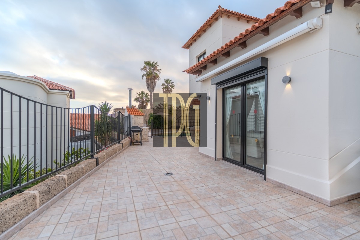 Terrace of a modern villa with sliding glass doors, tiled patio, and a black iron fence; palm trees and planters along the edge in the distance.