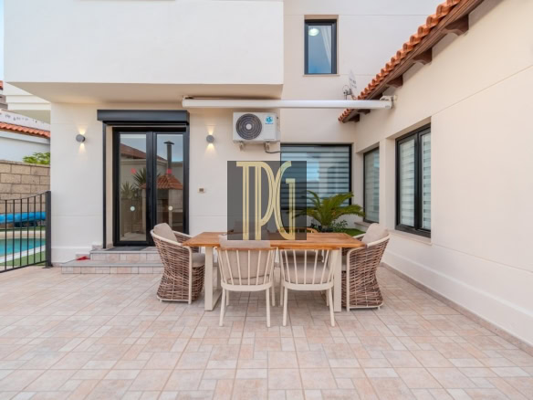 Outdoor patio of a modern home featuring a wooden dining table with six chairs on beige tiled floor, sliding glass doors behind, and a pool area visible to the left.