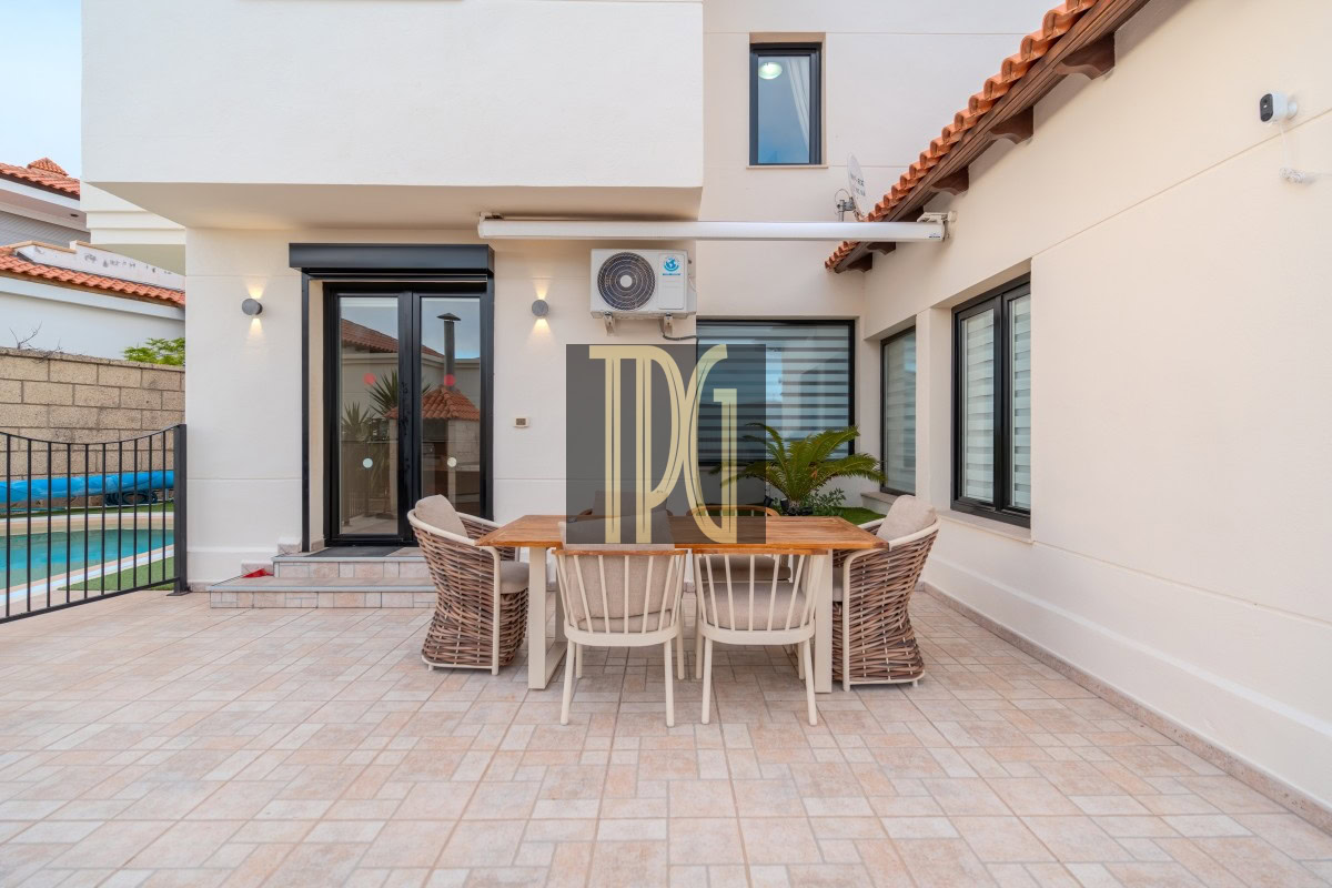 Outdoor patio of a modern home featuring a wooden dining table with six chairs on beige tiled floor, sliding glass doors behind, and a pool area visible to the left.