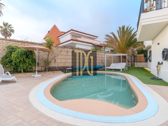 Kidney-shaped pool in a sunny backyard with beige tiled deck, a white lounge chair, umbrella, and palm trees along a privacy wall.