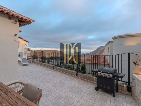 Terrace with tiled floor, black railing, and a barbecue grill, overlooking rooftops and distant mountains at sunset.