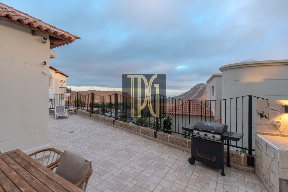 Terrace with tiled floor, black railing, and a barbecue grill, overlooking rooftops and distant mountains at sunset.