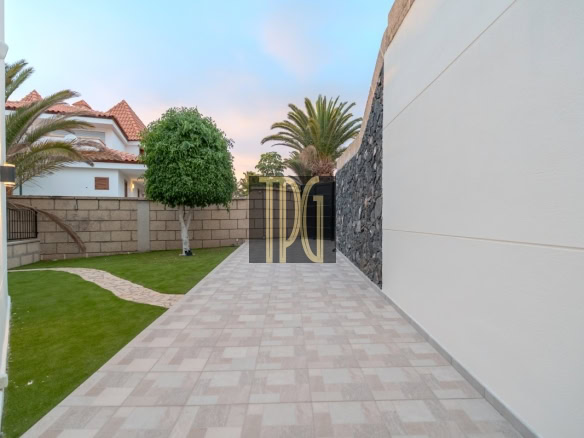 Wide tiled pathway in a modern backyard, bordered by green lawn, palm trees, and a stone wall on the right. Behind are white houses with red-tiled roofs.