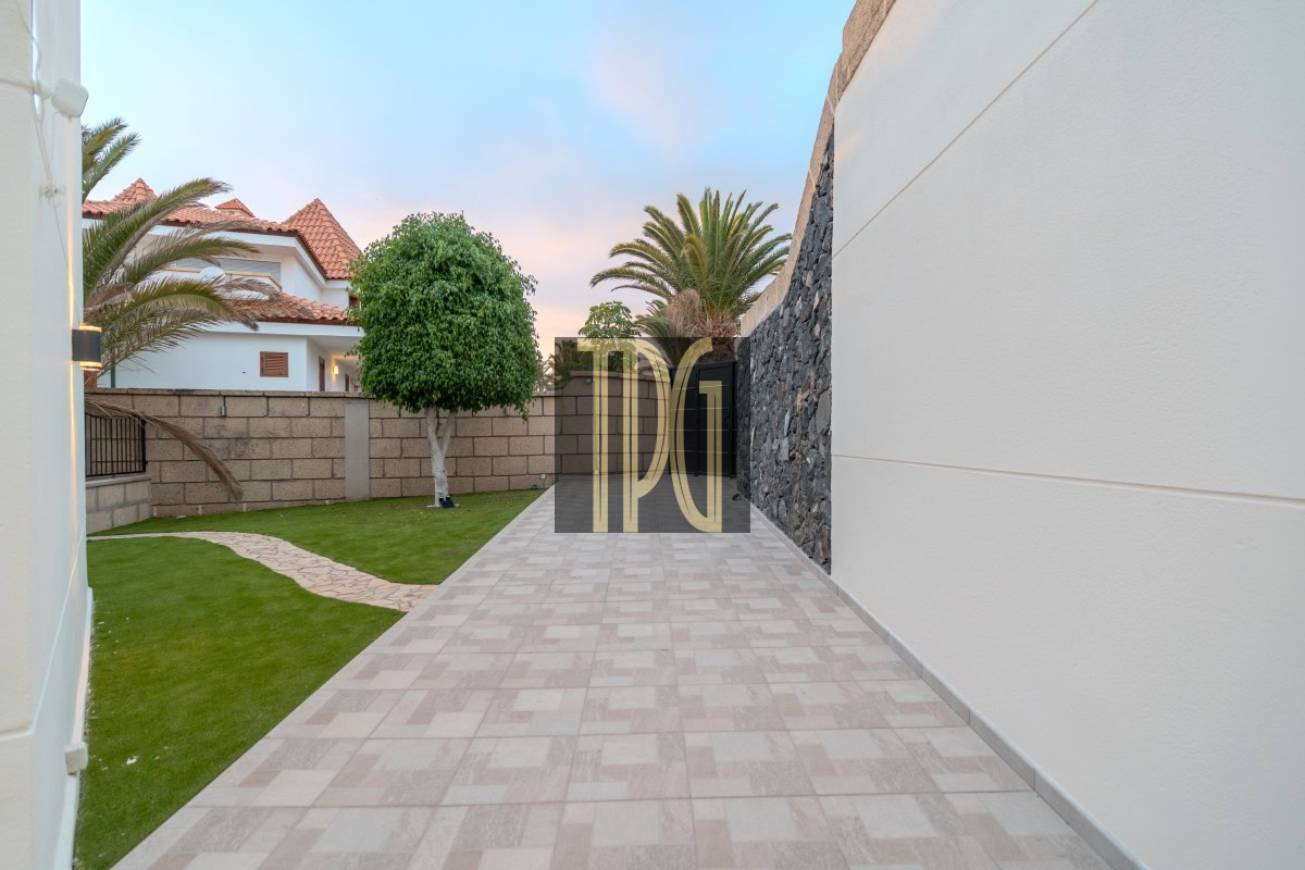 Wide tiled pathway in a modern backyard, bordered by green lawn, palm trees, and a stone wall on the right. Behind are white houses with red-tiled roofs.