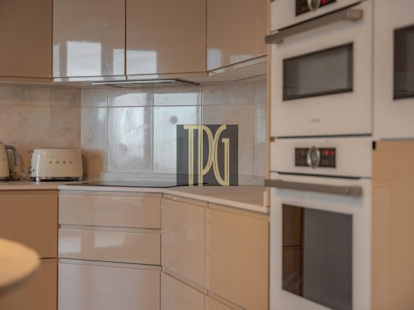 Beige modern kitchen with glossy cabinets, a white kettle and toaster on the counter, and built-in double ovens on the right.