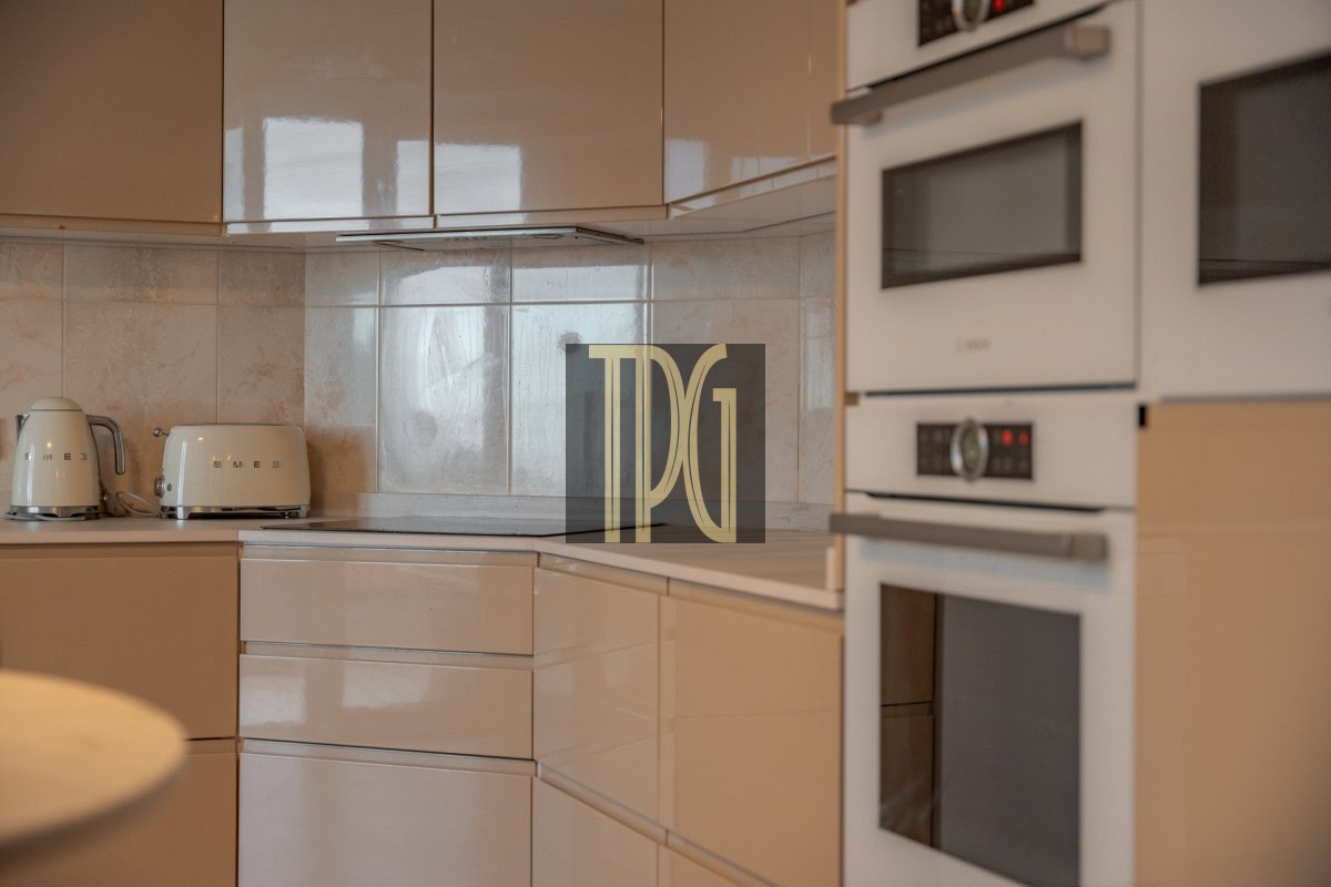 Beige modern kitchen with glossy cabinets, a white kettle and toaster on the counter, and built-in double ovens on the right.