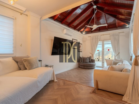 Bright living room with a white sofa set, beige armchairs, and a wooden vaulted ceiling with a ceiling fan and large windows leads to a balcony.